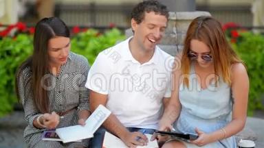 两名女学生与一个男孩一起学习户外坐在长凳上的城市公园。 电子学习概念。